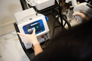 Fototapeta premium Top view of beauty therapist hands holding laser resurfacing machine, pointing with finger at the monitor screen, adjusting programs on hardware beauty apparatus