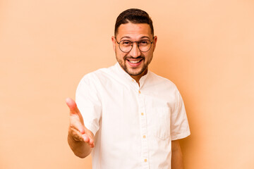 Young hispanic man isolated on beige background stretching hand at camera in greeting gesture.