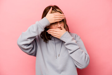 Young caucasian woman isolated on pink background blink at the camera through fingers, embarrassed covering face.