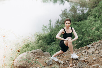 Naklejka na ściany i meble Young female runner stretching arms and legs before running at morning forest trail near lake