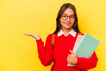 Young student hispanic woman isolated on yellow background showing a copy space on a palm and holding another hand on waist.