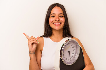Young hispanic woman holding a scale isolated on white background smiling and pointing aside,...