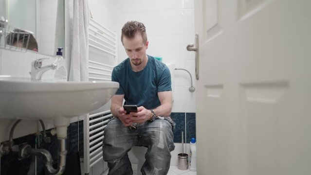 Open door to bathroom with man on toilet using smartphone 4K