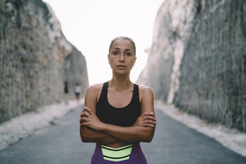 Half length of Caucaisan female runner with crossed hands keeping healthy lifestyle posing at road between rocks, muscular fit girl in tracksuit looking at camera during sportive photo session