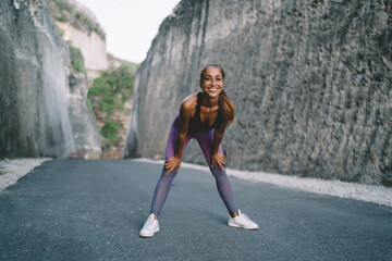 Portrait of cheerful woman in active wear feeling happy during morning training - smiling at camera, joyful Caucaisan female in sportive clothes posing at road between rocks enjoying healthy lifestyle