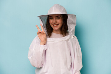 Young caucasian beekeeper woman isolated on blue background joyful and carefree showing a peace symbol with fingers.