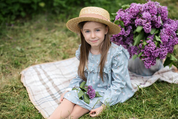 Beautiful little girl in dress with Lilac bouquet in garden. Spring blossom. Cute Smile young girl  in straw hat with purple flowers on summer picnic. Gardening. Adorable child outdoors. childhood