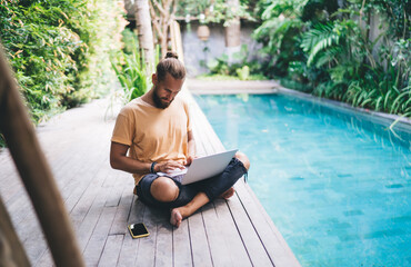 Caucasian man watching browsed video on modern netbook computer working remotely at pool terrace,...
