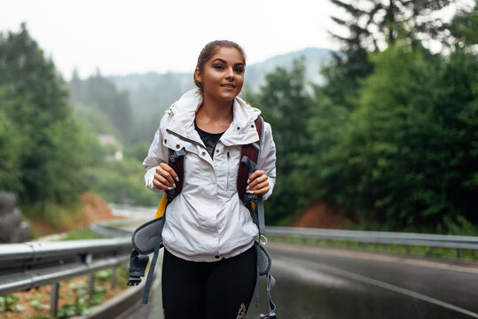 Happy young woman with backpack walking on wet road and enjoying beautiful view on mountains