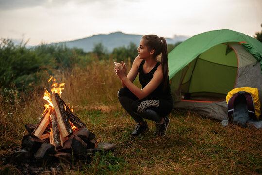 Woman squatting near green tent and warming hands over bonfire