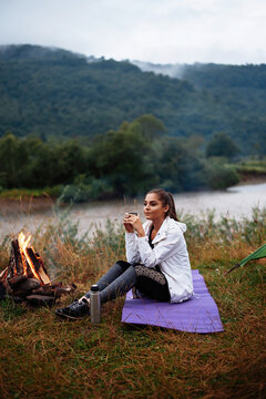 Young girl sitting near camping fire and drinking tea