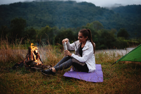 Woman pouring tea from thermos bottle near burning fire outdoors