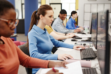 Vibrant side view shot of college students using computers in row, focus on Caucasian young woman doing research in library