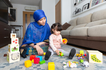 Young Muslim woman playing with her young daughter in the living room. Single parent family at home. 