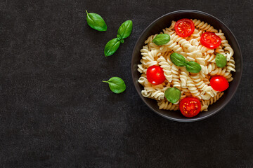 Fusilli pasta cooked with tomatoes and basil leaf, on dark background, top view, space to copy text.