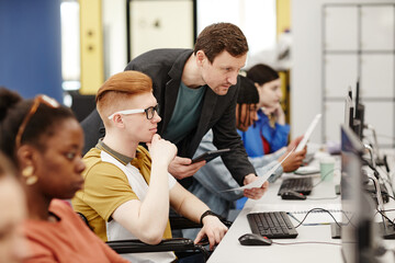 Vibrant side view portrait of male teacher helping student using computer in college classroom,...