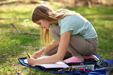 A young adolescent girl writes or colors in a notebook or journal on a blanket on the grass 