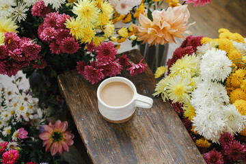 Warm cup of coffee and beautiful autumn flowers on rustic wooden background. Good morning. Stylish mug with cappuccino among colorful asters and dahlias composition. Moody image