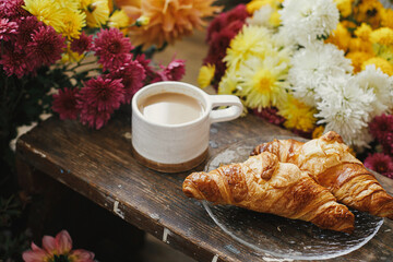 Warm cup of coffee and freshly baked croissants on rustic wooden background with beautiful autumn flowers. Good morning. Stylish mug with cappuccino and croissants on plate, moody image