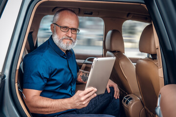 Shot of elderly businessperson with tablet looking at camera inside of car in daytime.