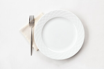 Empty white plate with fabric napkin and a fork on white table. Top view.