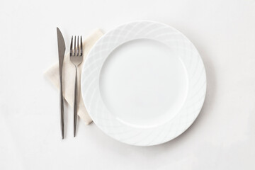 Empty white plate with fabric napkin, a fork and a knife on white table. Top view.