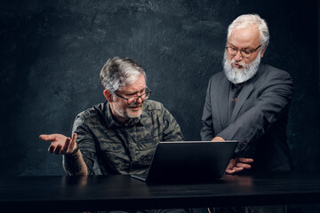 Shot of two old men with laptop talking and discussing some ideas against dark background.