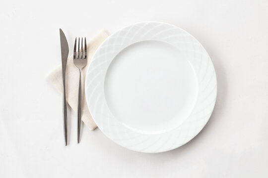 Empty white plate with fabric napkin, a fork and a knife on white table. Top view.
