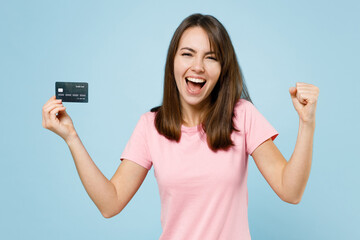 Young happy cool fun woman 20s wear pink t-shirt look camera hold in hand credit bank card do winner gesture isolated on pastel plain light blue background studio portrait. People lifestyle concept.