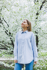 Young pretty woman enjoys standing near flowering spring tree. A girl wearing beige hat and white dress smiles among blooming apple trees. Spring season concept