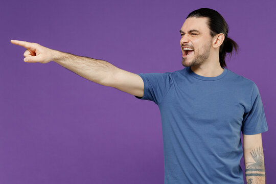 Young angry furious sad abuser man 20s wearing basic blue t-shirt point index finger aside scream shout say do it isolated on plain purple color background studio portrait. People lifestyle concept.