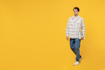 Full body young smiling happy cheerful caucasian man 20s wear white casual shirt hold closed laptop pc computer walk go isolated on plain yellow background studio portrait. People lifestyle concept.