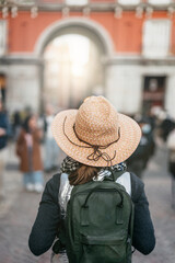 a woman hiker, with her back turned, walks along an old square in Madrid. she is wearing a straw hat and carrying a backpack. concept of vacations and tourism in spain.