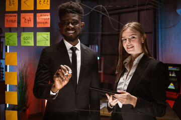 Sharing of business ideas. Two multiracial people in formal wear using adhesive notes and writing while standing behind the glass wall in the board room. Work in the evening office