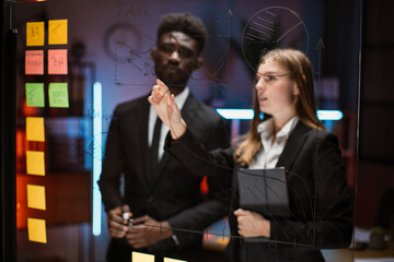 View through the office glass board. Blur view of two young focused mixed race financiers or economists, discussing project and sharing ideas, working near kanban glass board with sticky notes
