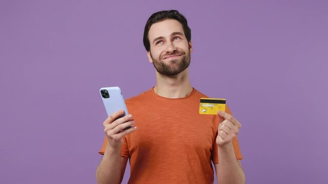 Dreamful happy young brunet bearded man 20s in red t-shirt using mobile cell phone hold in hand credit bank card doing online shopping isolated on plain pastel light purple background studio portrait