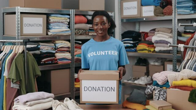 Portrait view of the multiracial volunteer woman holding box with clothes for donation and looking at the camera with smile. Shelves with belongings at the background. Donations concept