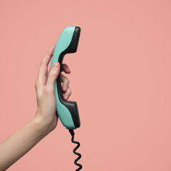 A woman's hand holds a blue retro telephone receiver. Pink background.