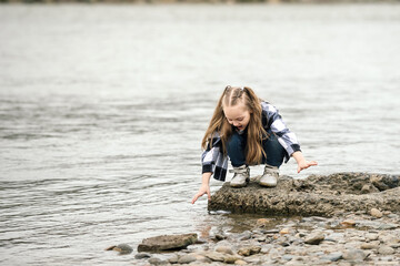 a girl with blond long hair, in a fashionable plaid shirt and jeans, has fun by the river on a walk during school holidays