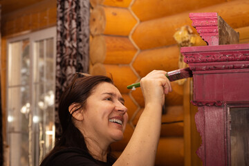Happy woman with smile painting wooden ornamented cupboard in pink with paint brush with window in...