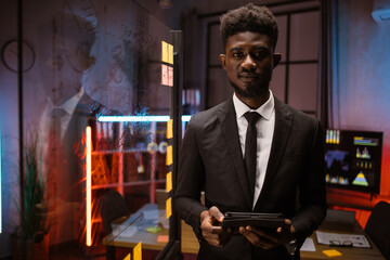 Agile methodology, business concept. Portrait of young handsome smart African American man in formal clothes, standing with tablet pc near glass kanban board with sticky notes at dark office