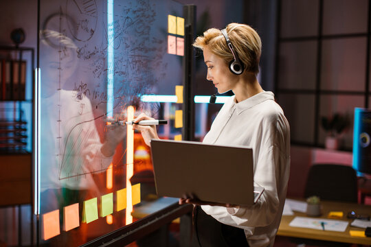 Female financial expert, ceo manager working near scrum kanban glass wall and writing ideas. Blond woman in headset, using laptop and consulting with colleague on video call