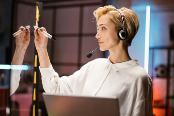 Close up side view of confident blond young businesswoman wearing white shirt and headset, writing...