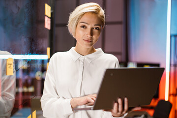 Smart focused blond businesslady, wearing white shirt, typing on laptop while standing near...