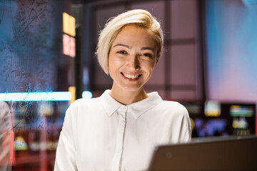 Close up head shot of pretty smiling blond Caucasian business woman in white shirt, posing on camera, while standing in modern dark office. Blurred office interior background, glass board