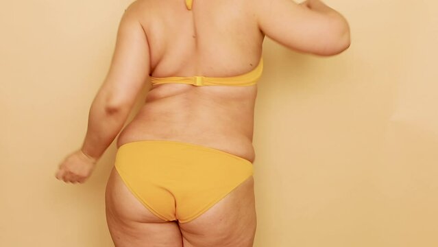 Back view of unrecognizable fat plump woman wearing yellow bra, bikini, dancing, making active moves, waving hands, moving buttocks on beige background. Body positive, dance, weight loss, having fun.
