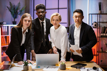 Portrait of creative multiethnic business team standing together at the table with gadgets and papers and looking at camera. Multiracial business people together at the evening meeting.