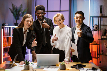 Portrait of creative multiethnic business team standing together at the table with gadgets and papers and looking at camera. Multiracial business people together at the evening meeting.