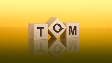 Three wooden cubes with the letters TQM on the bright yellow surface. the inscription on the cubes is reflected from the gray surface of the table. TOM - Total Quality Management acronym