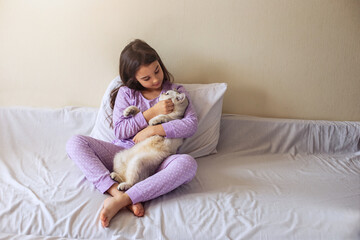 A little girl in purple pajamas, with her dark hair loose, sits hugging a white charming cat